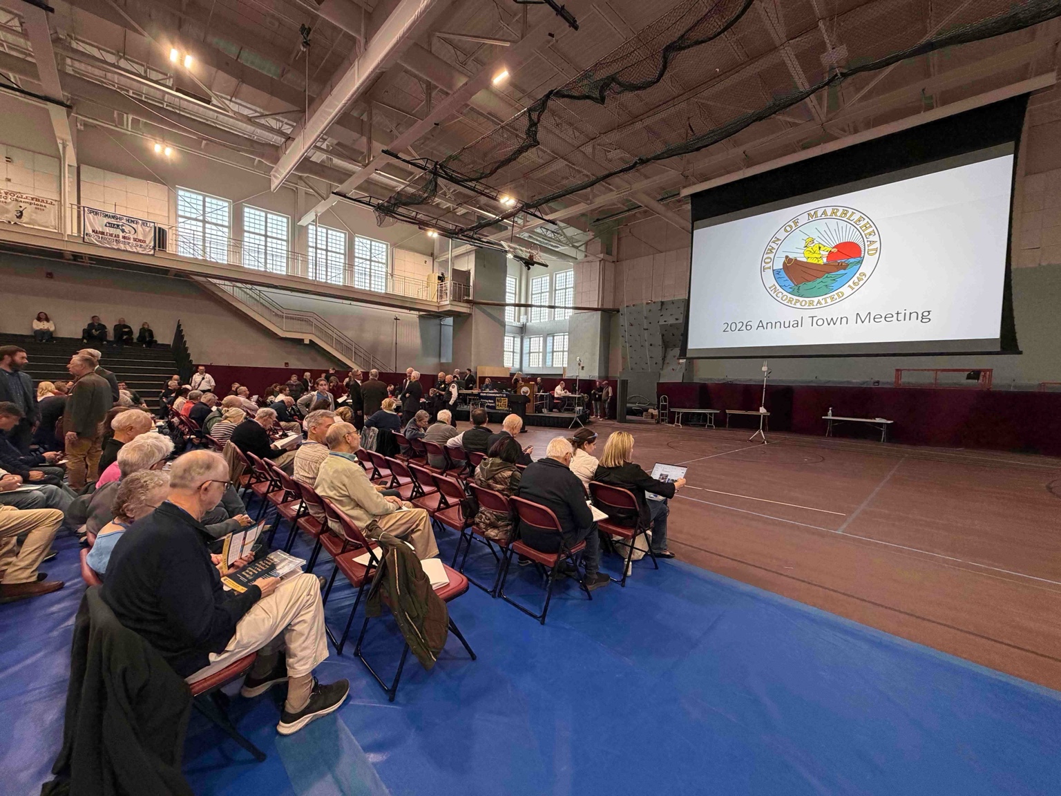 Residents settle into red folding chairs across the gym floor as Town Meeting begins.