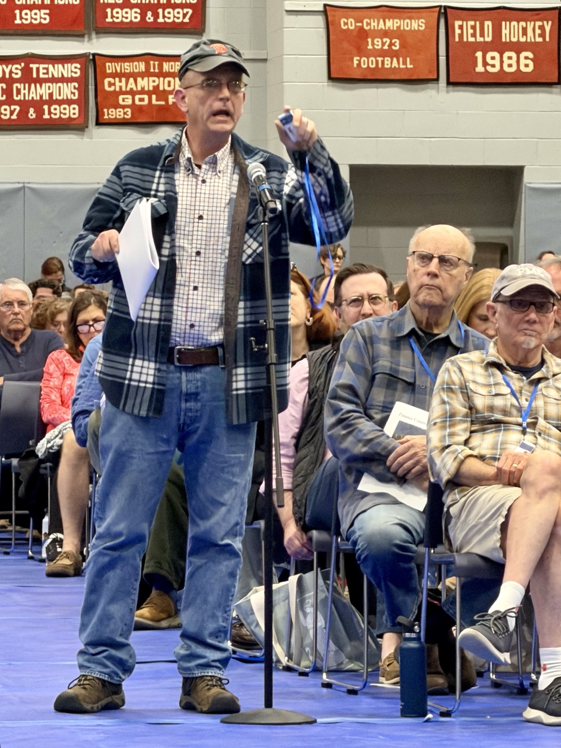 James Full at the aisle microphone, holding up his Town Meeting clicker on its blue lanyard as he challenges the moderator's reliance on hand votes.