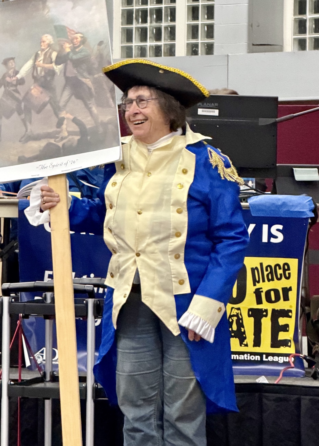 Lynn Nadeau in a Continental Army officer's coat and tricorn hat holds a print of The Spirit of '76 as she presents Article 40 on the floor of Town Meeting.