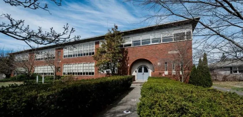 Long two-story brick school building with an arched entry, hedges and bare trees