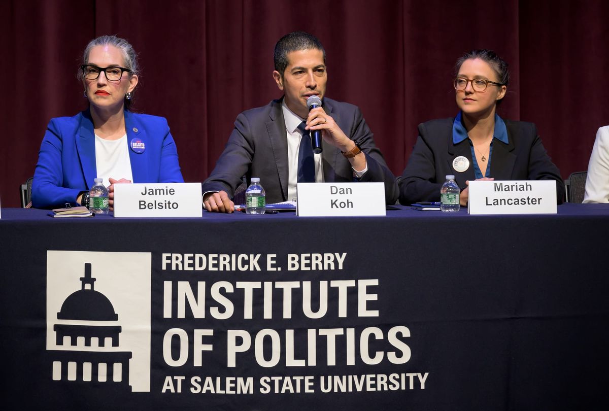 Three candidates at a debate table with nameplates, the man at center speaking into a microphone