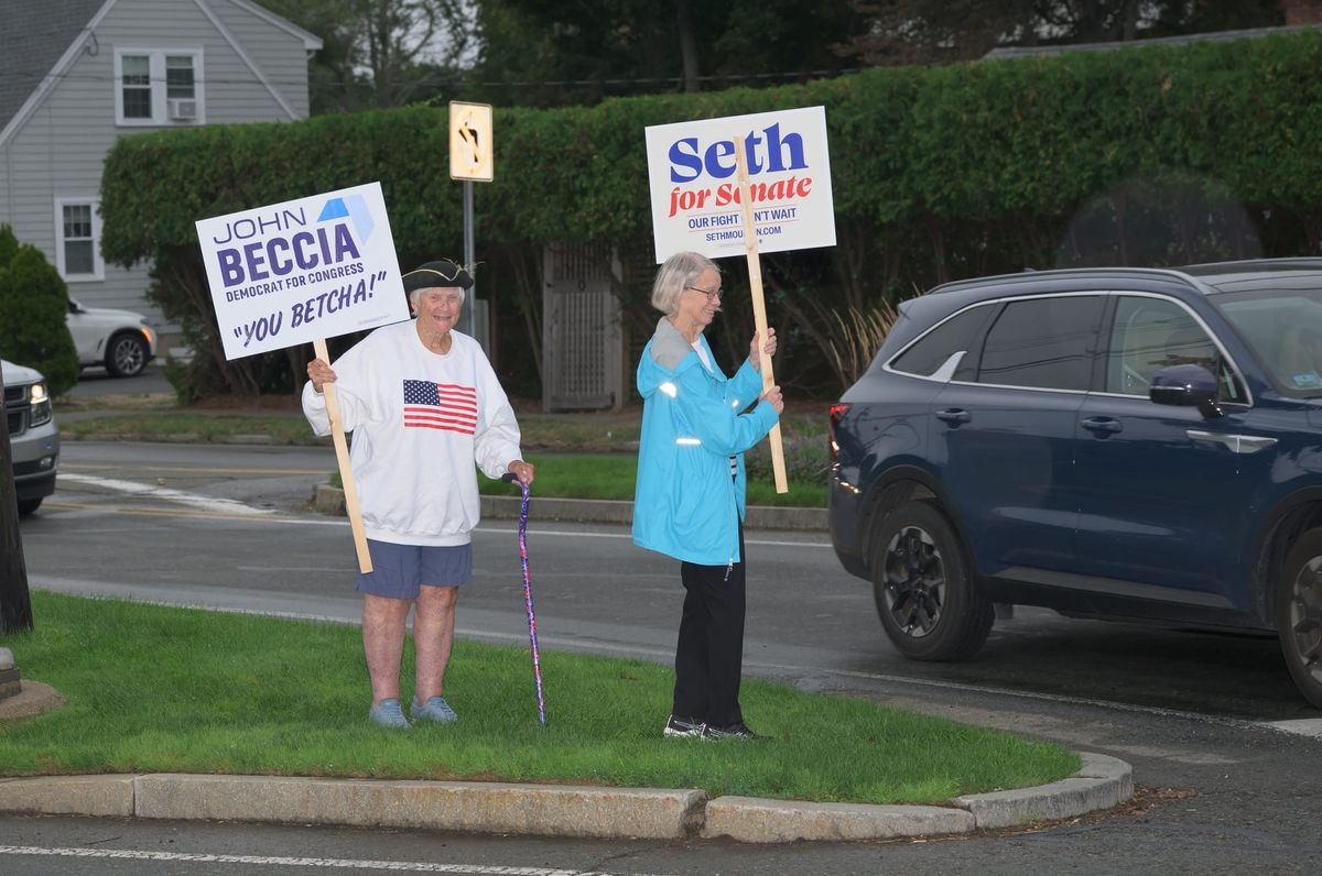 Two older women on a grass median holding campaign signs beside a roadway