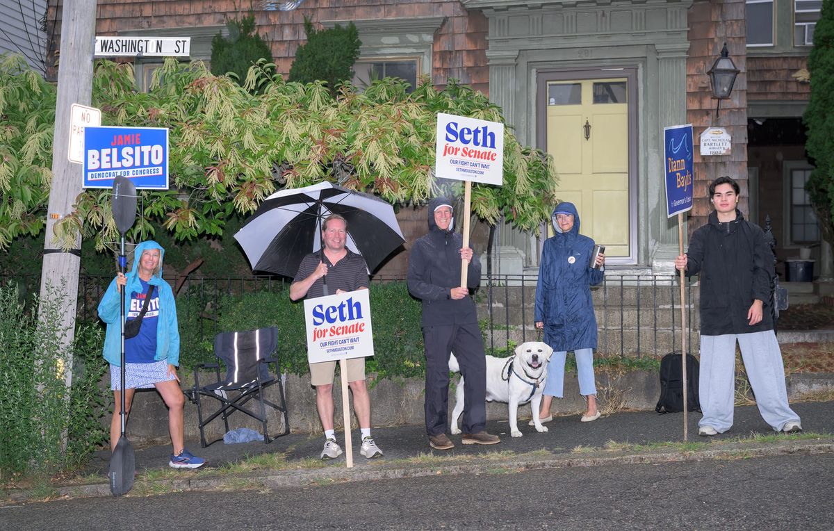Five people in rain gear holding campaign signs on a sidewalk with a dog
