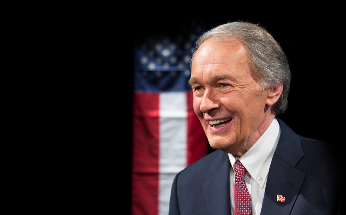 Smiling white-haired man in a dark suit and red tie before a blurred American flag